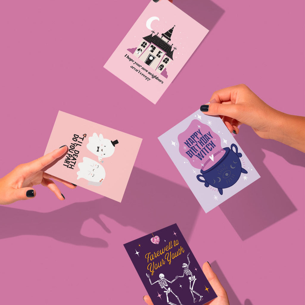 Four Halloween-themed greeting cards held by hands on a pink background